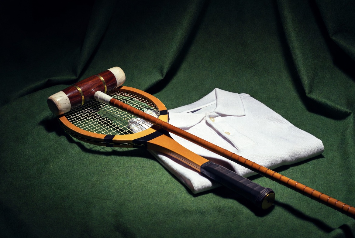 Cinematic still life of a vintage tennis racket crossed with a polo mallet resting on a folded white petit piqué polo shirt, dramatic lighting on deep green felt — symbolizing the collision of French court performance and American equestrian heritage.